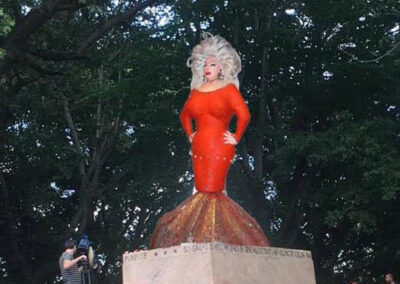 Photo of Divine in a red fishtail dress standing on a pedestal in a park.